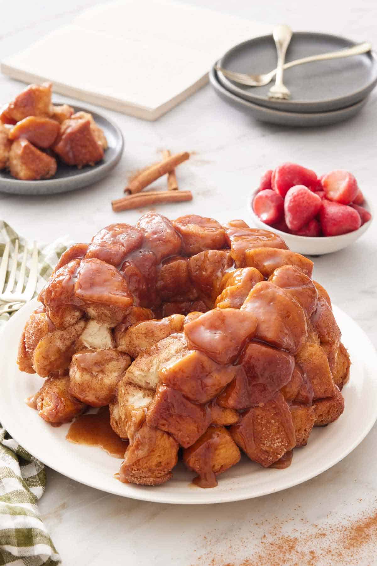 A platter with monkey bread with a slice cut out. Plated serving, strawberries, cinnamon sticks, stack of bowls, forks, and a notebook in the background.