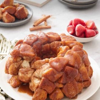 Pinterest graphic of a platter with monkey bread with a slice cut out. Plated serving, strawberries, cinnamon sticks, stack of bowls, forks, and a notebook in the background.