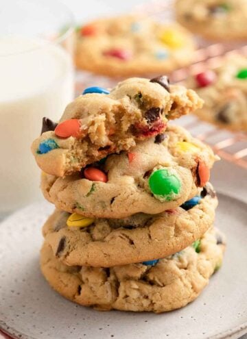 A stack of three and a half monster cookies on a plate. Glass of milk in the background and more cookies.