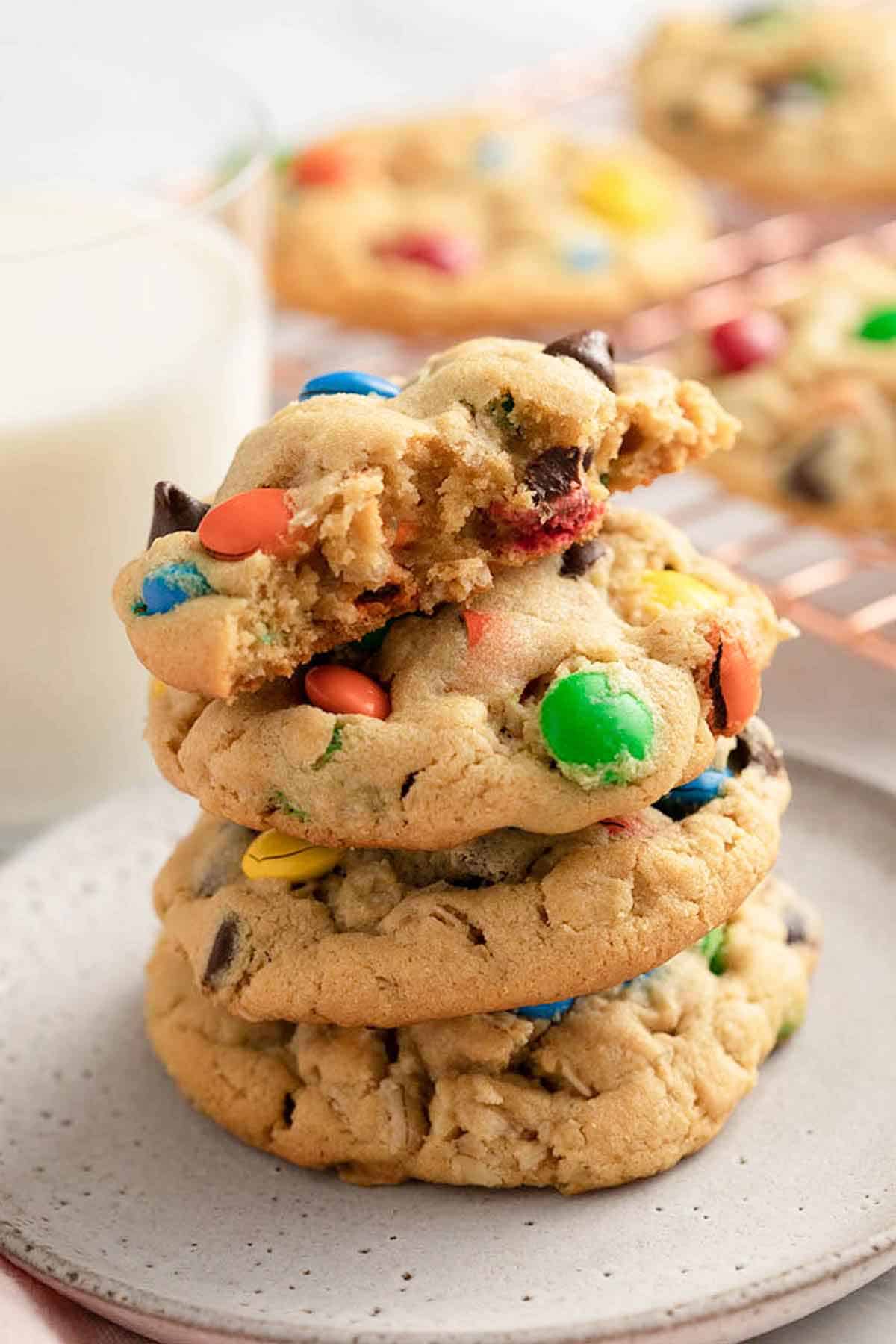 A stack of three and a half monster cookies on a plate. Glass of milk in the background and more cookies.