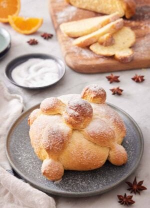 A loaf of Pan de Muerto with a wooden serving board in the back with sliced bread.