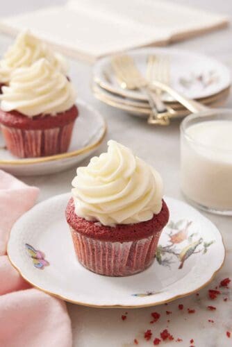 A red velvet cupcake with a glass of milk in the back along with a few more cupcakes, plates, and forks.