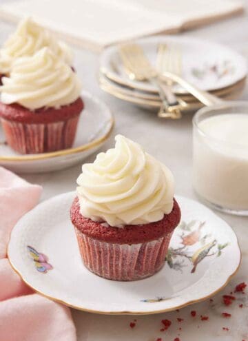 A red velvet cupcake with a glass of milk in the back along with a few more cupcakes, plates, and forks.