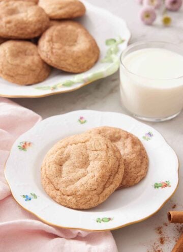 A plate with two snickerdoodle cookies with a glass of milk and additional cookies in the background.