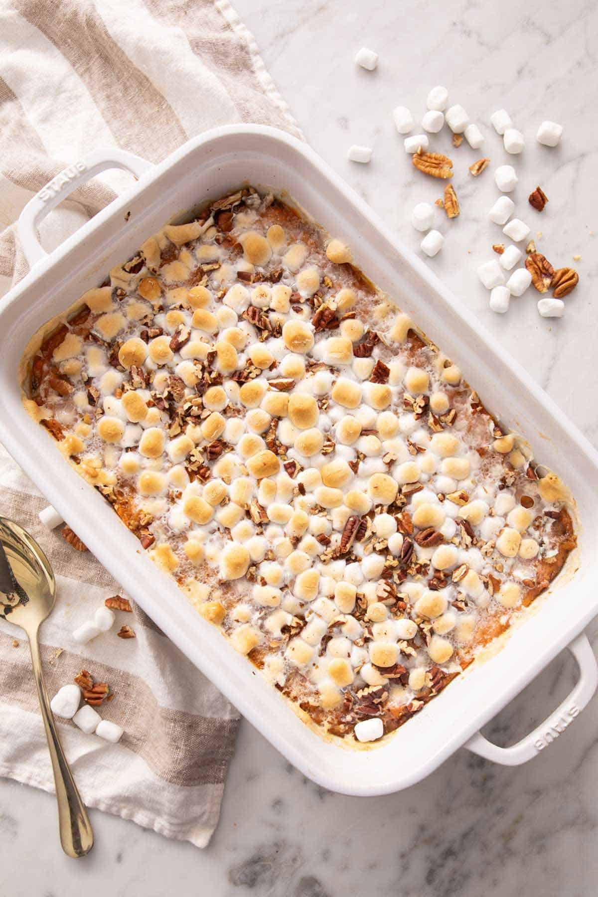 An overhead view of a baking dish of sweet potato casserole with marshmallows and pecans scattered around with a linen and spoon beside it.