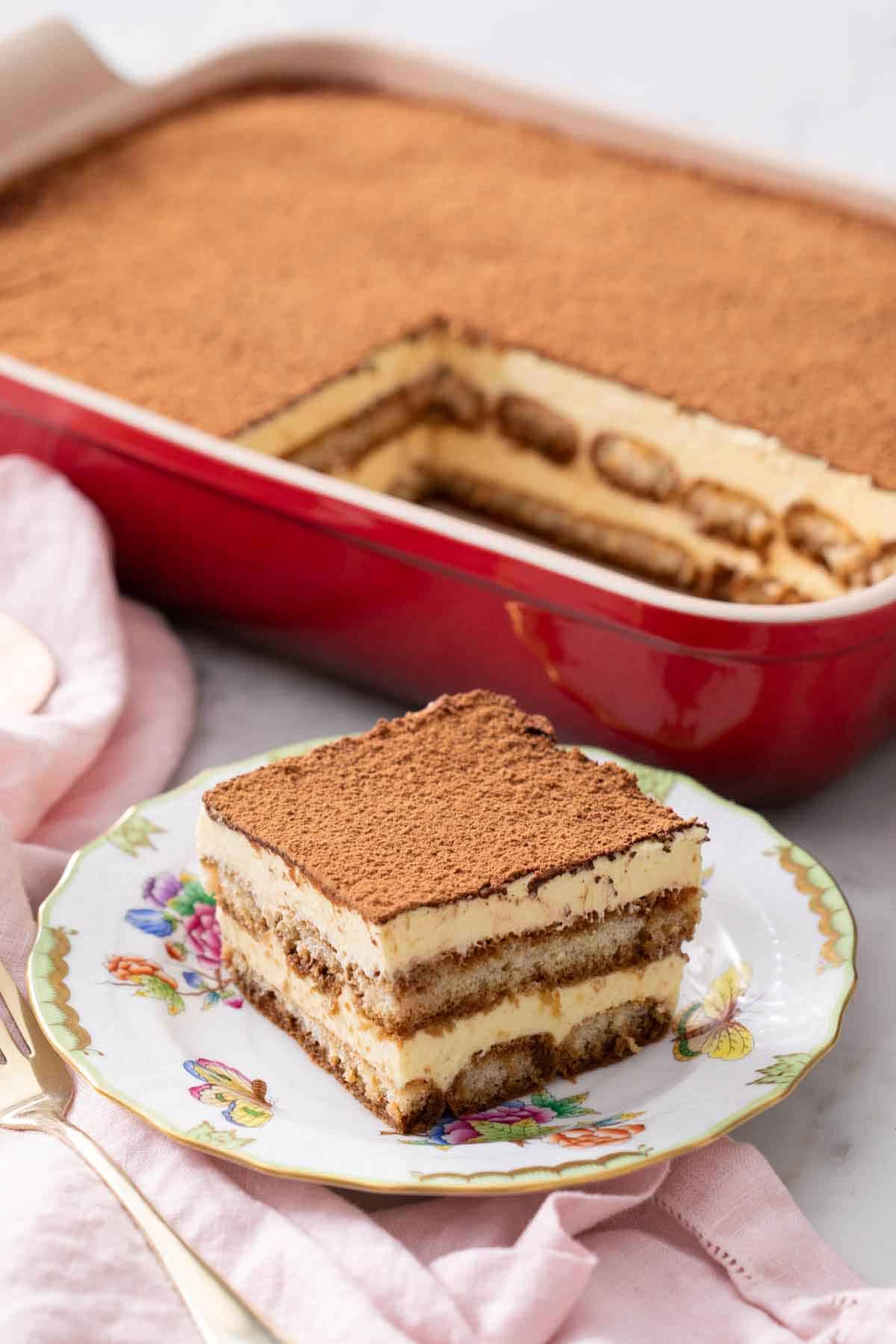A serving of tiramisu on a plate with the rest in a baking dish in the background.