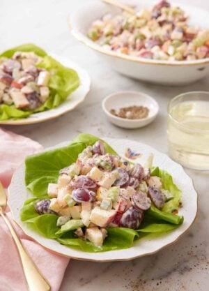 A plate with Waldorf salad on a lettuce leaf with another plated serving and a platter of salad in the background. A glass of wine and some pepper behind the plate.