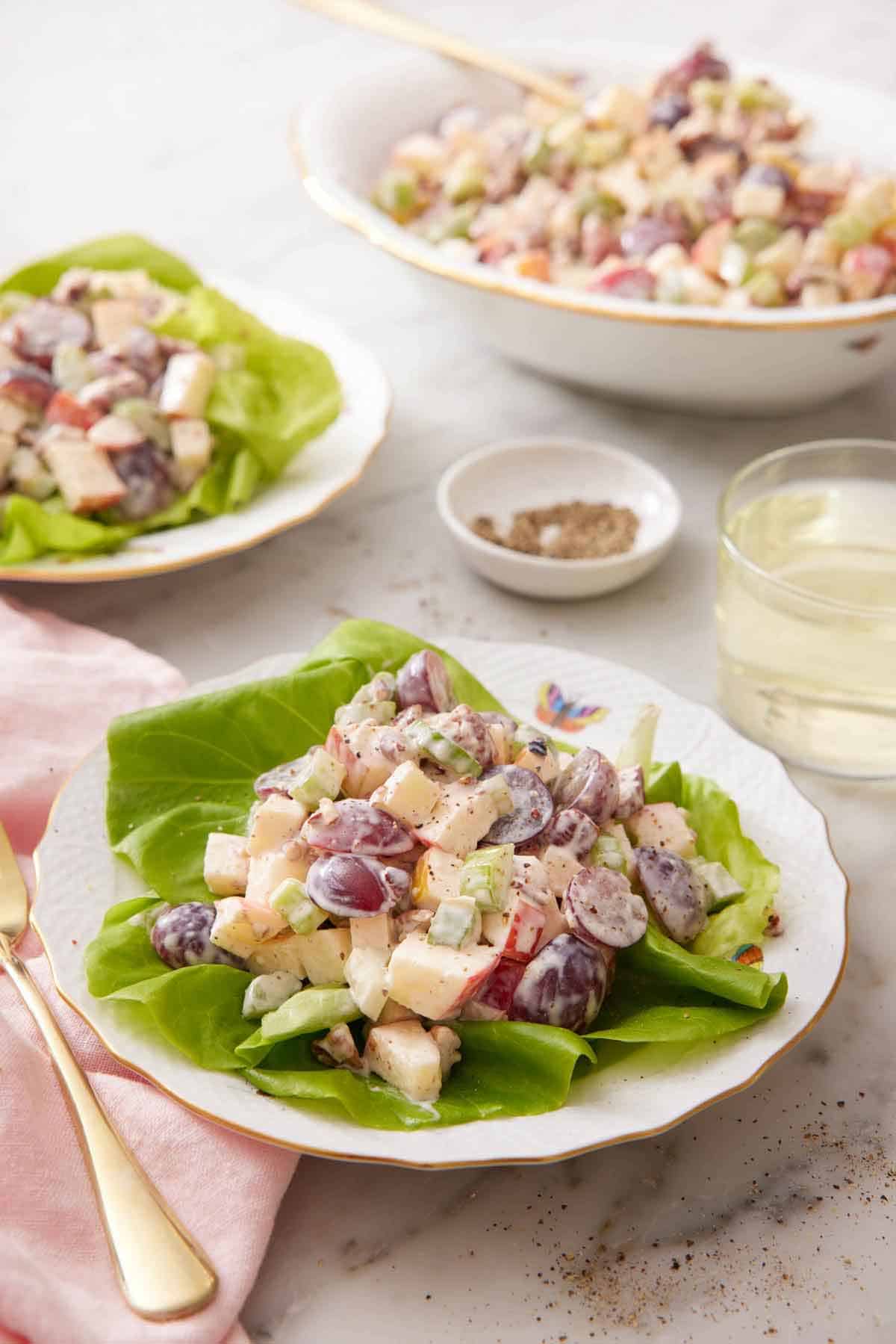 A plate with Waldorf salad on a lettuce leaf with another plated serving and a platter of salad in the background. A glass of wine and some pepper behind the plate.