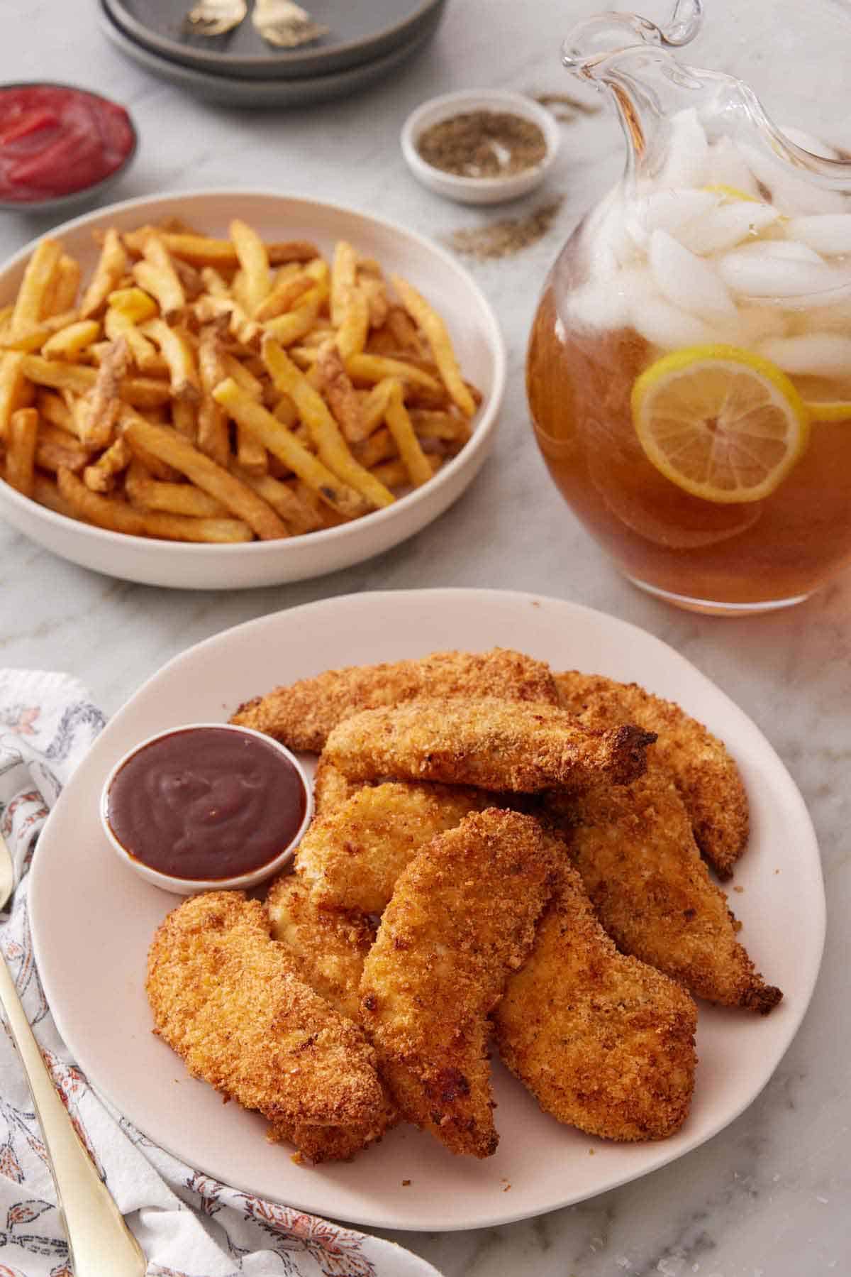 A plate of air fryer chicken tenders with a small bowl of bbq sauce. A bowl of french fries and a pitcher of iced tea in the background.