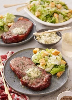 A plate with an air fryer steak with a side of salad. An additional plated serving in the background along with a platter of salad.