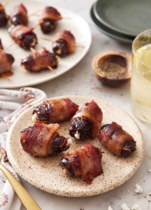 A small plate with five bacon wrapped dates with a platter in the background along with pepper and a drink.