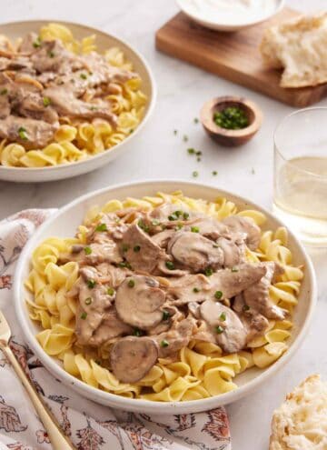 A plate of beef stroganoff topped with chives with a second plate, a drink, and torn bread in the background.