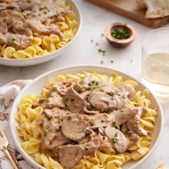 Pinterest graphic of a plate of beef stroganoff topped with chives with another plate, a drink, and torn bread in the background.