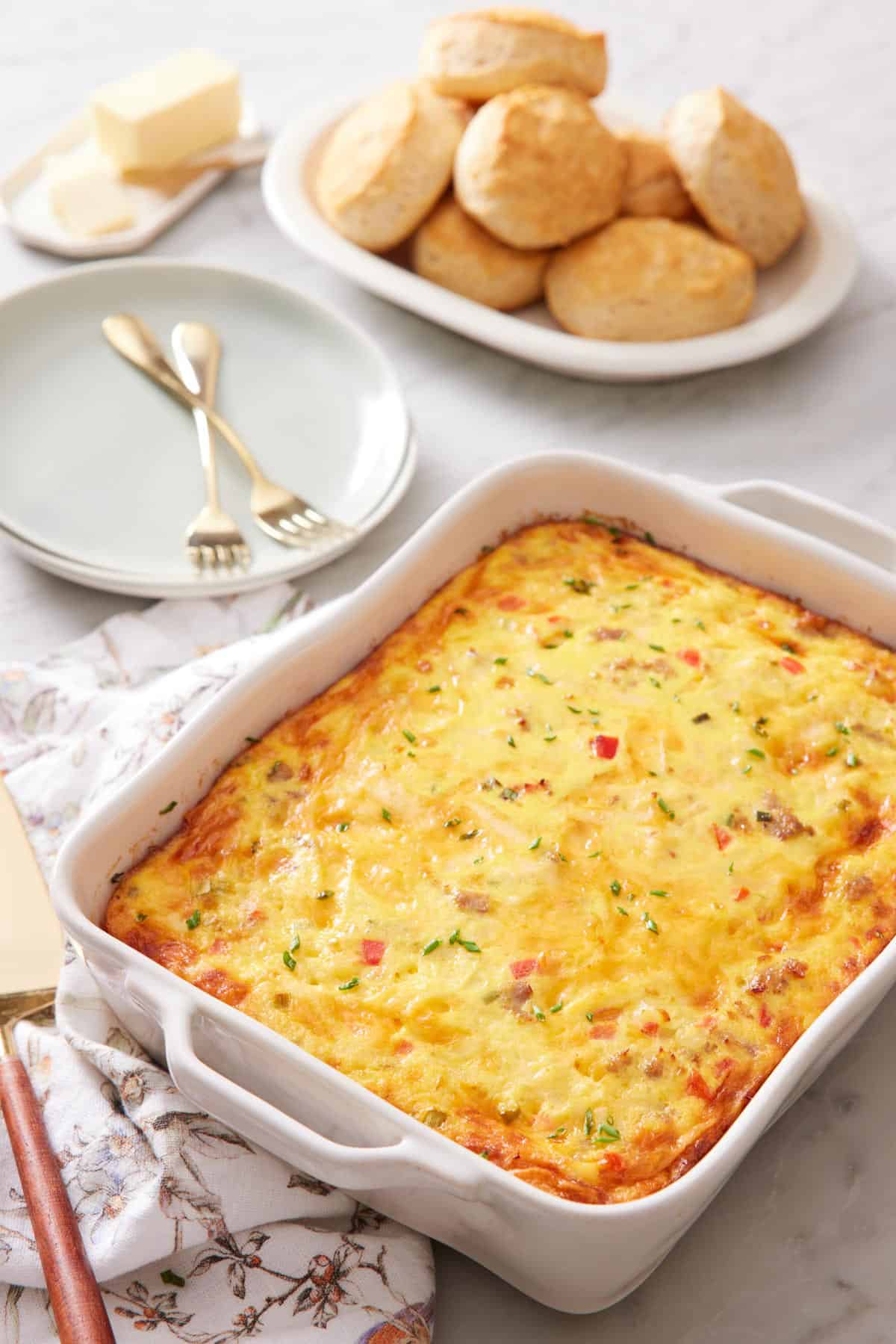 A baking dish of breakfast casserole with a plate of rolls and stack of plates with forks in the background.