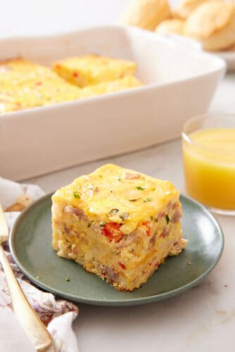 A plate with a serving of breakfast casserole with a baking dish and orange juice in the background.