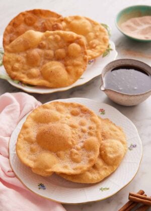 A plate with two pieces of bunuelos with more in the background along with the piloncillo syrup.