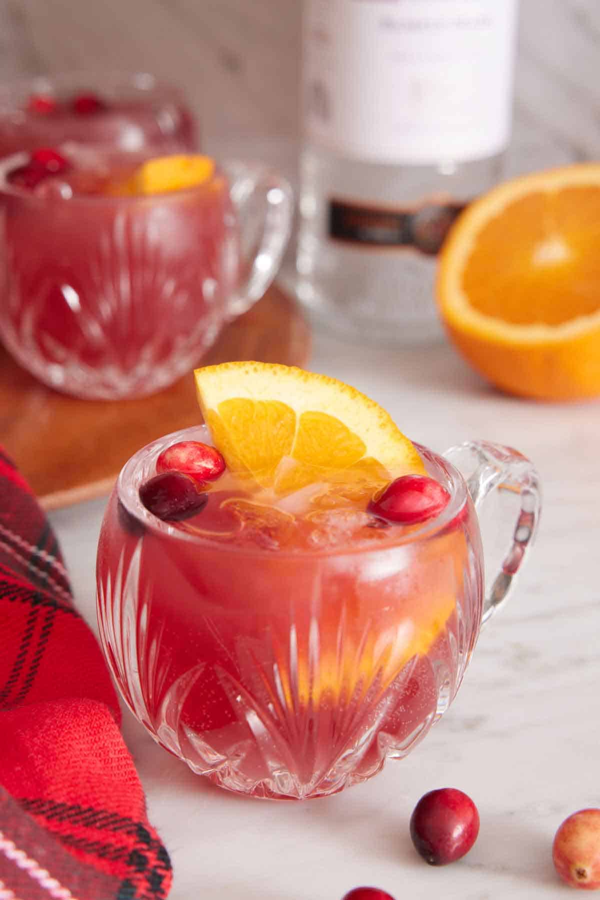 A glass of Christmas punch with orange and cranberries with more in the background along with a cut orange and bottle of vodka.