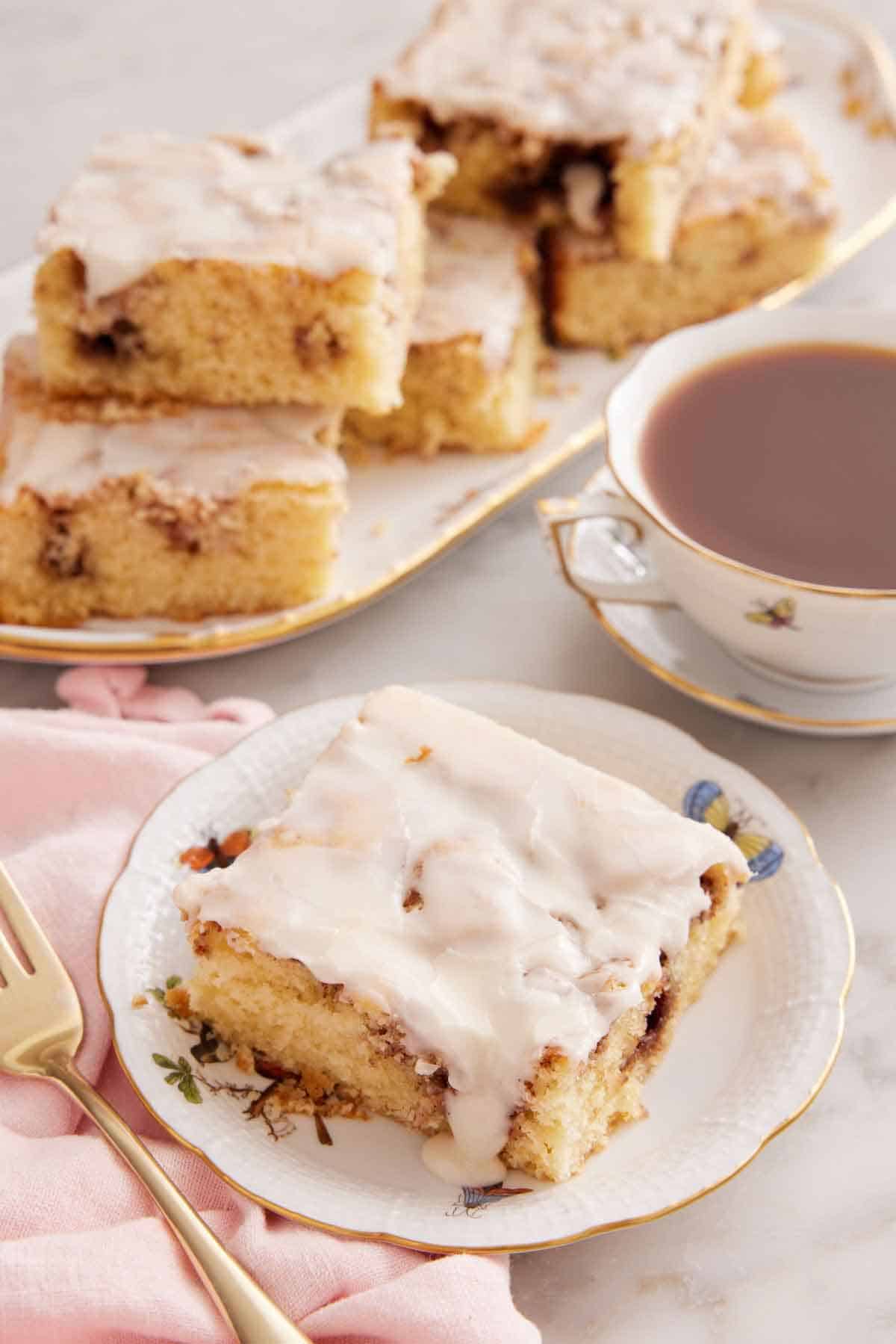 A plate with a slice of cinnamon roll cake with a mug of coffee and platter of cut cake in the background.