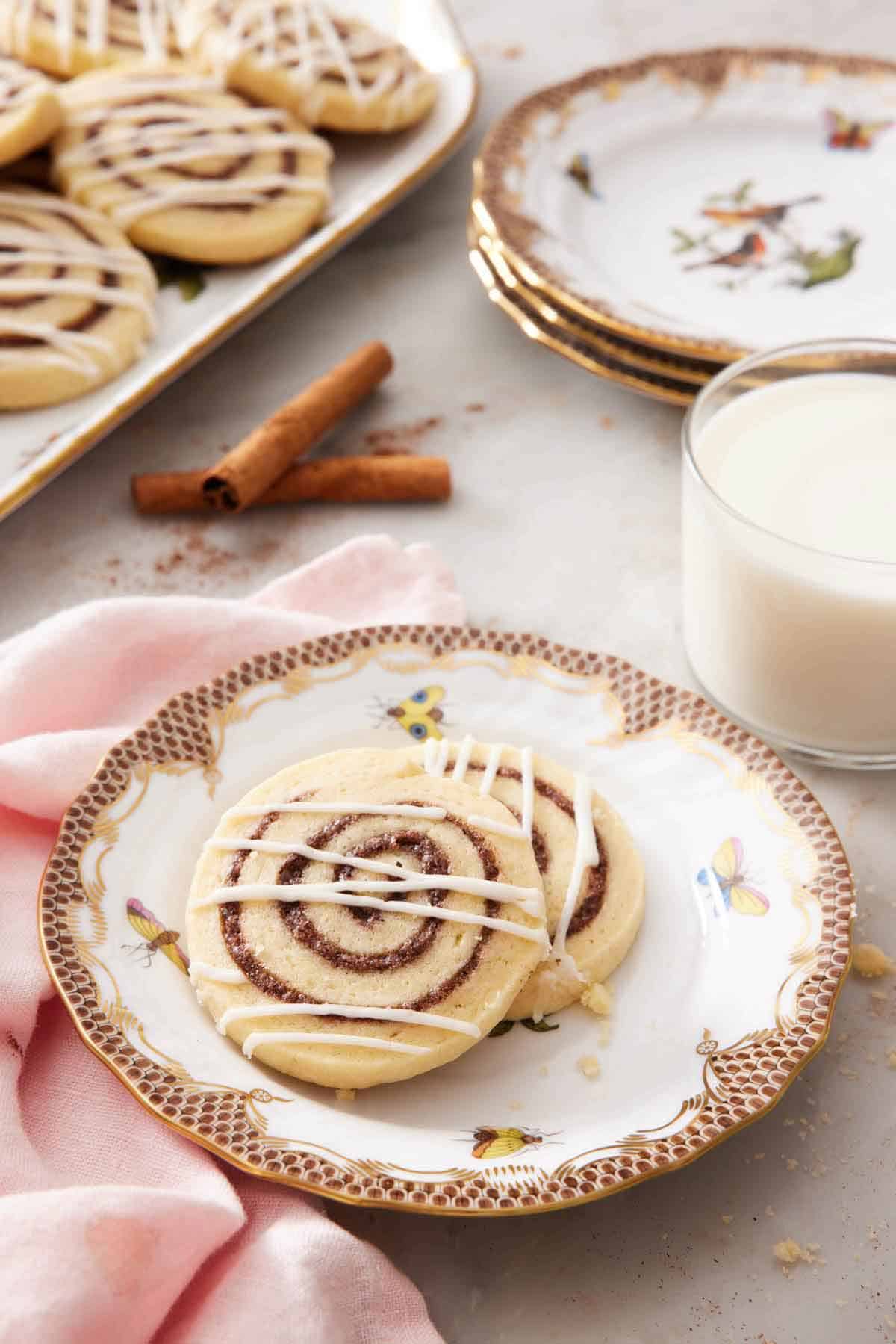 A plate with two cinnamon roll cookies along with a glass of milk, stack of plates, and more cookies in the background.