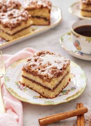 A plate with a slice of coffee cake with some cinnamon sticks in front and cup of coffee with more cake in the background.