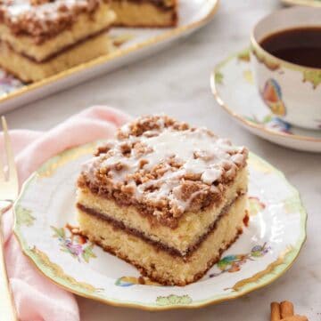 A plate with a slice of coffee cake with some cinnamon sticks in front and cup of coffee with more cake in the background.