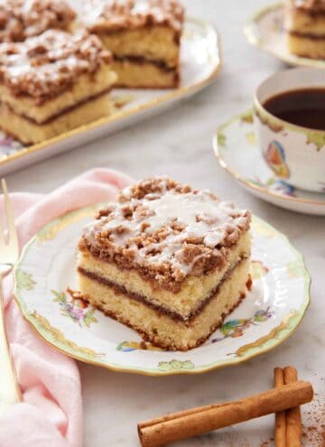 A plate with a slice of coffee cake with some cinnamon sticks in front and cup of coffee with more cake in the background.