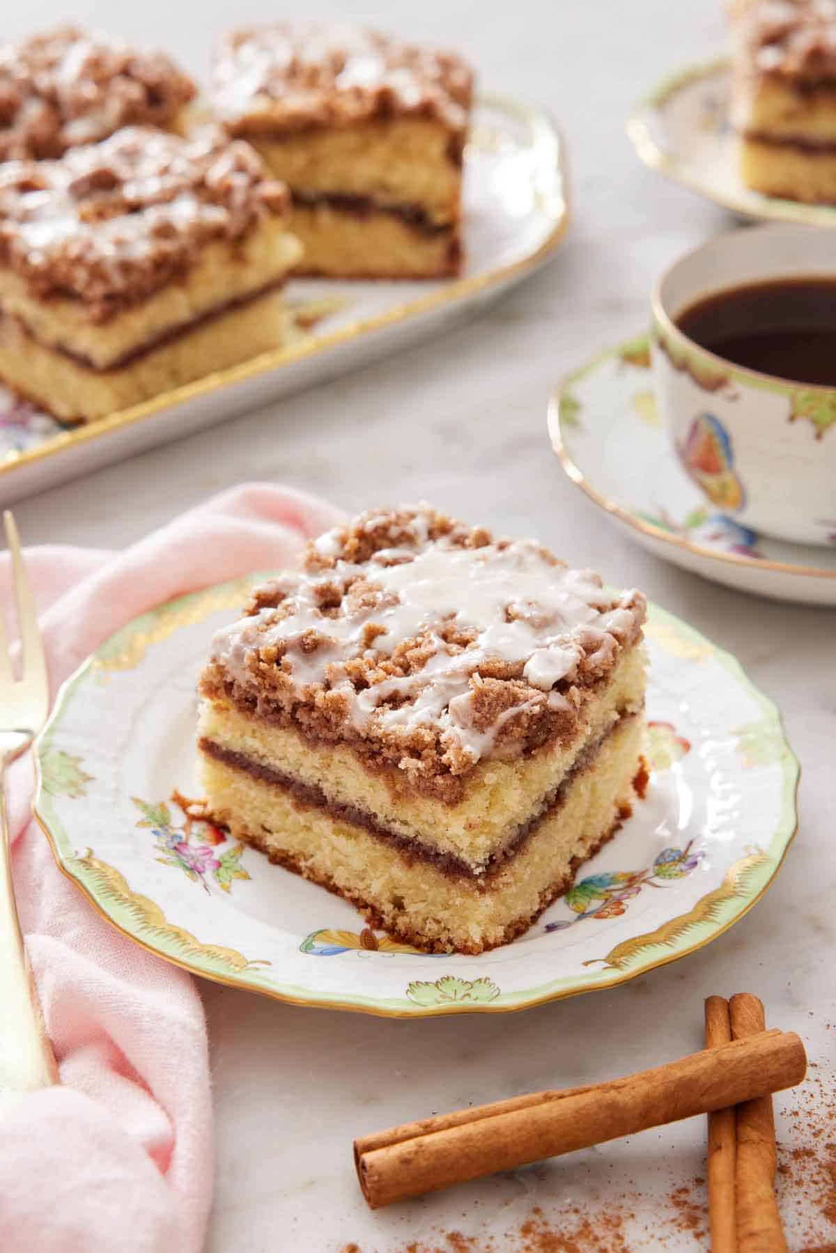 A plate with a slice of coffee cake with some cinnamon sticks in front and cup of coffee with more cake in the background.