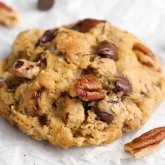 A cowboy cookie on a wrinkled parchment paper with chocolate chips and pecans scattered around it.
