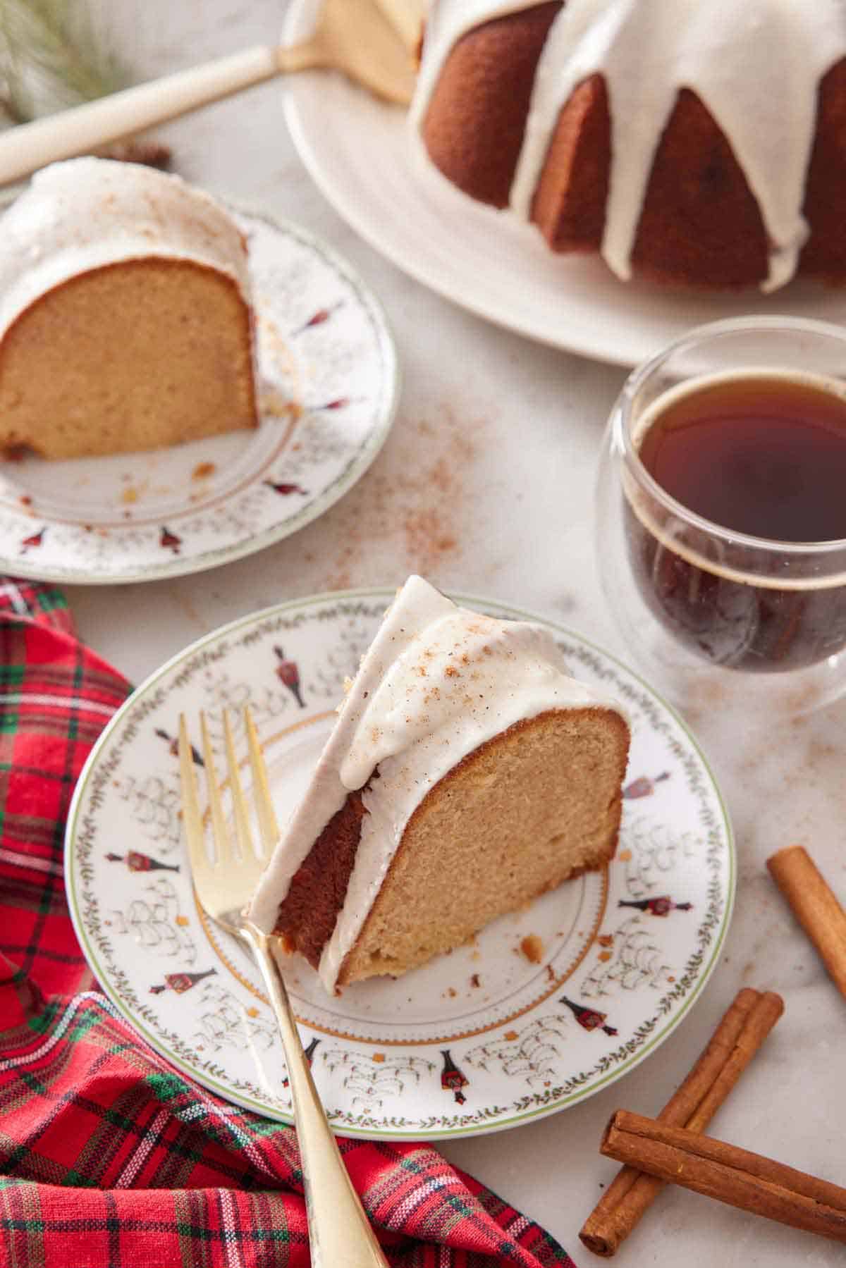 A festive plate with a slice of eggnog cake with a fork on the plate. Cinnamon sticks around it along with another plated slice, a drink, and the rest of the cake.