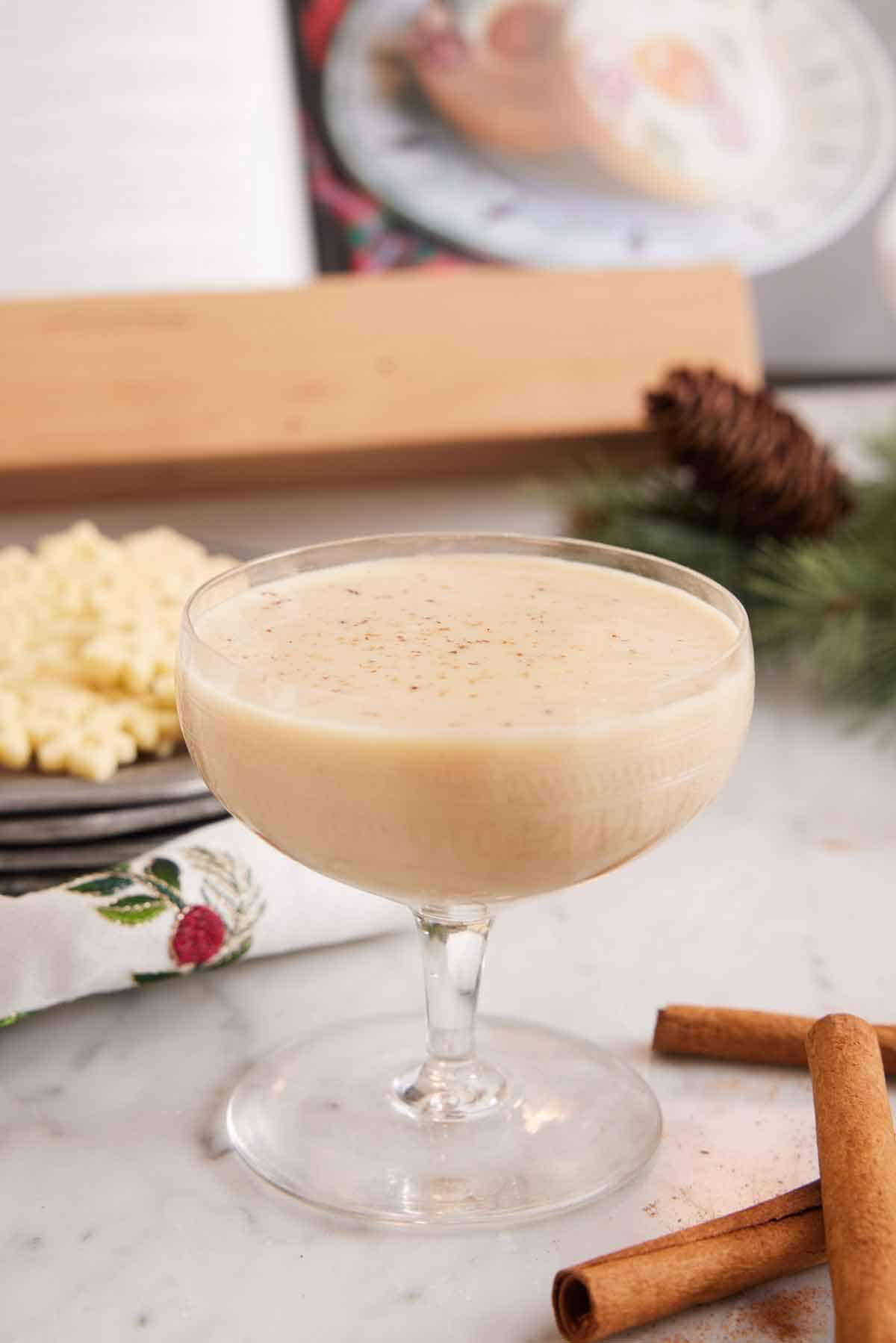 A glass of eggnog with some cinnamon sticks in the front and some cookies and pinecones in the background.