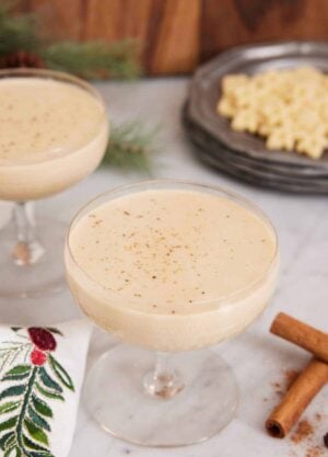 Two glasses of eggnog with one front and center. Cinnamon sticks in the front.
