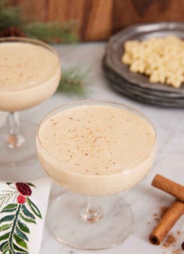 Two glasses of eggnog with one front and center. Cinnamon sticks in the front.