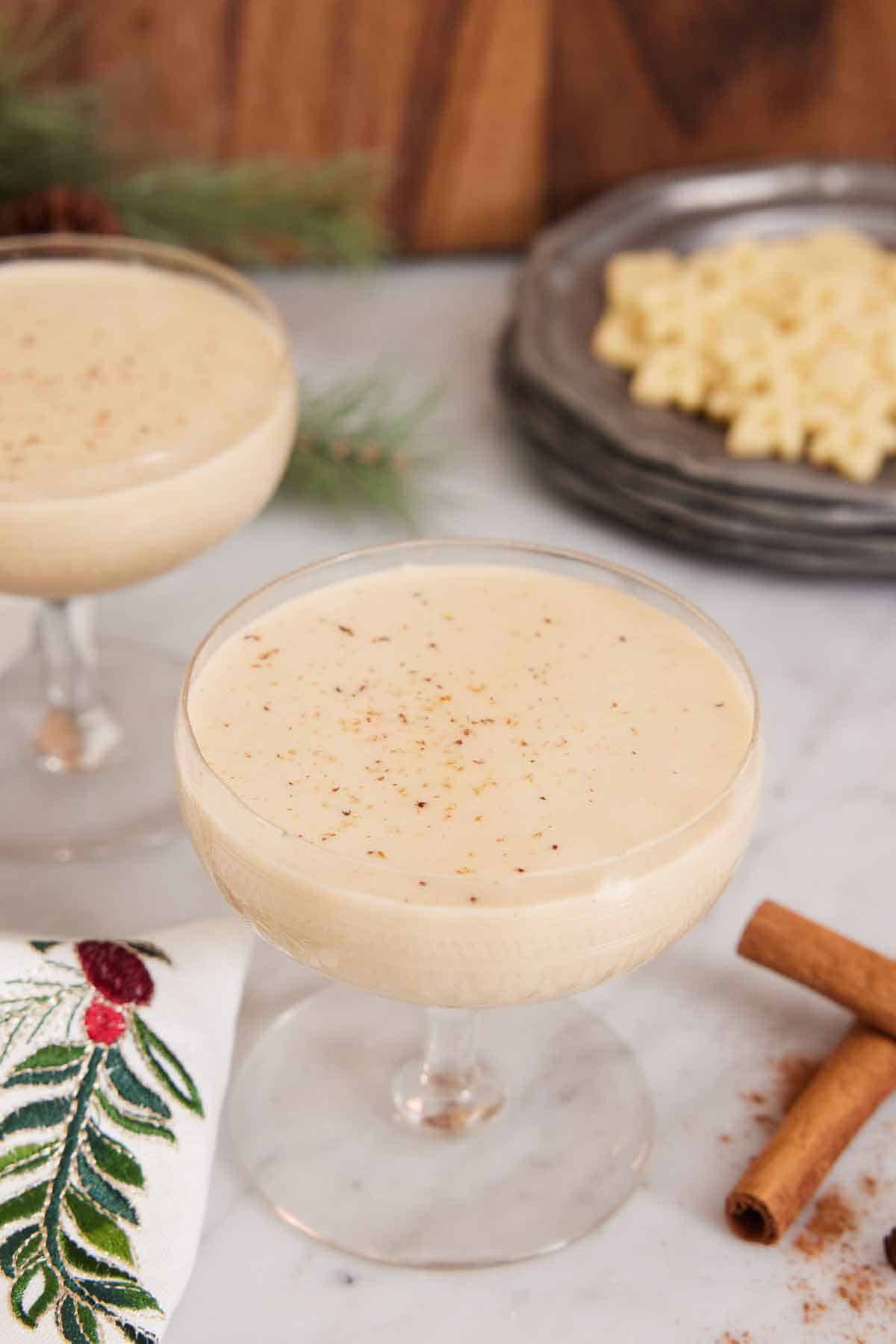 Two glasses of eggnog with one front and center. Cinnamon sticks in the front.