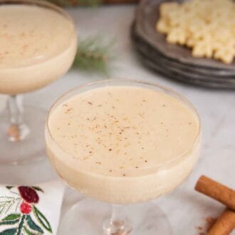 Pinterest graphic of two glasses of eggnog with one front and center. Cinnamon sticks in the front and cookies in the back.