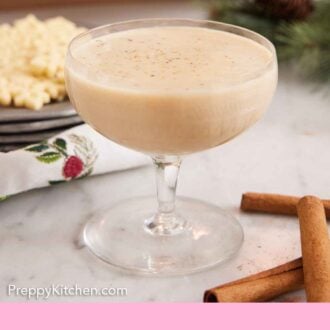 Pinterest graphic of a glass of eggnog with three cinnamon sticks in the front and some cookies and pinecones in the background.