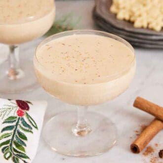 A glass of eggnog with a sprinkling of cinnamon on top. Cinnamon sticks on the side and cookies in the back.