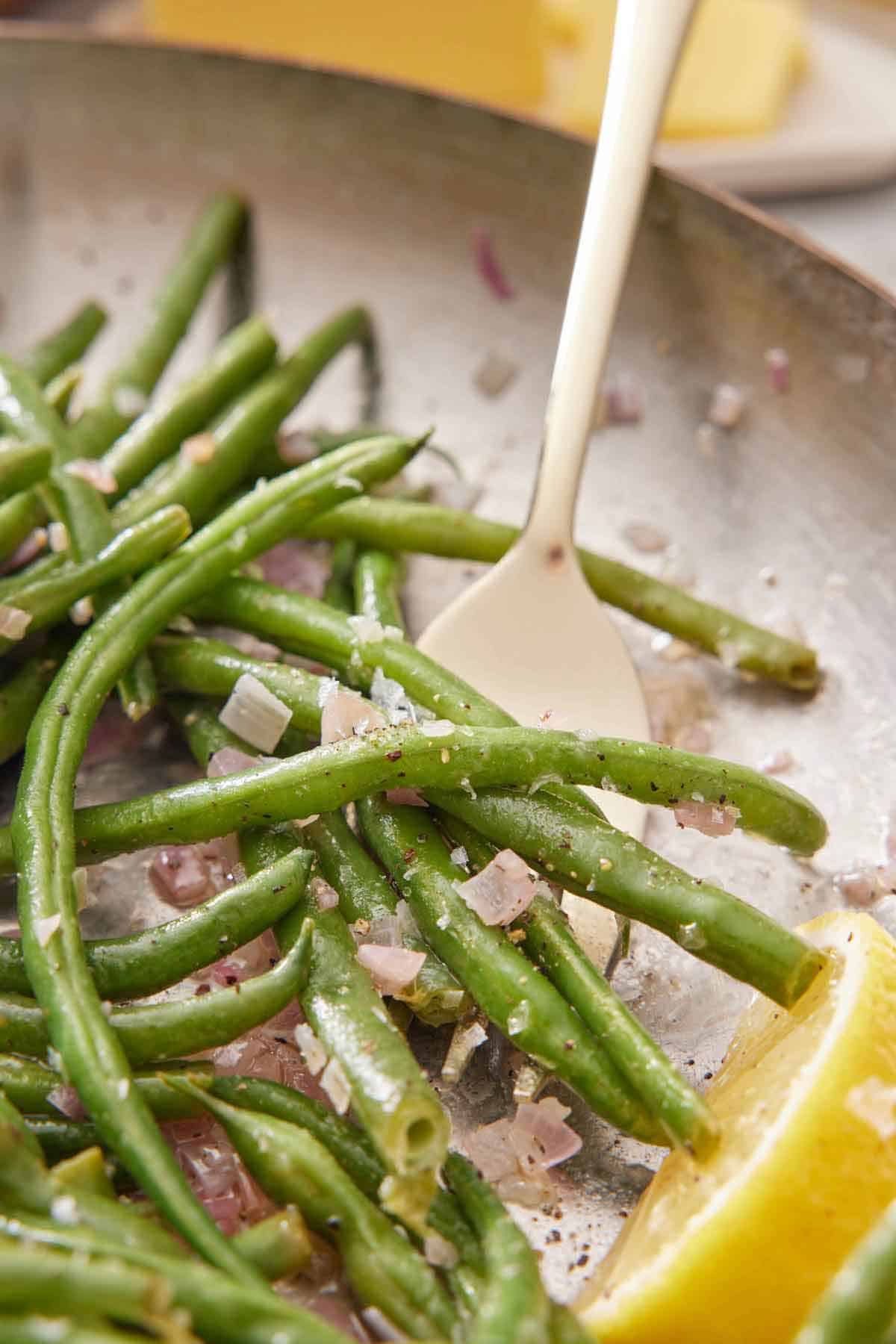 A skillet of French green beans with a fork underneath with a wedge of lemon beside it.