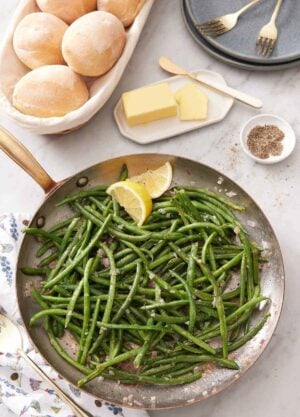 Overhead view of a skillet of French green beans with a side of dinner rolls, butter, and a bowl of pepper.