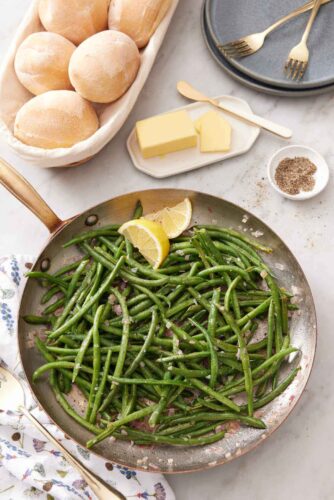 Overhead view of a skillet of French green beans with a side of dinner rolls, butter, and a bowl of pepper.