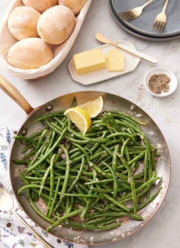 Overhead view of a skillet of French green beans with a side of dinner rolls, butter, and a bowl of pepper.
