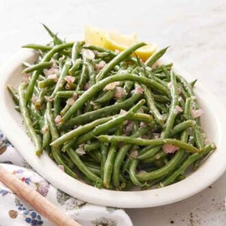 A platter of French green beans surrounded with a wooden spoon, stack of plates, forks, and some pepper.