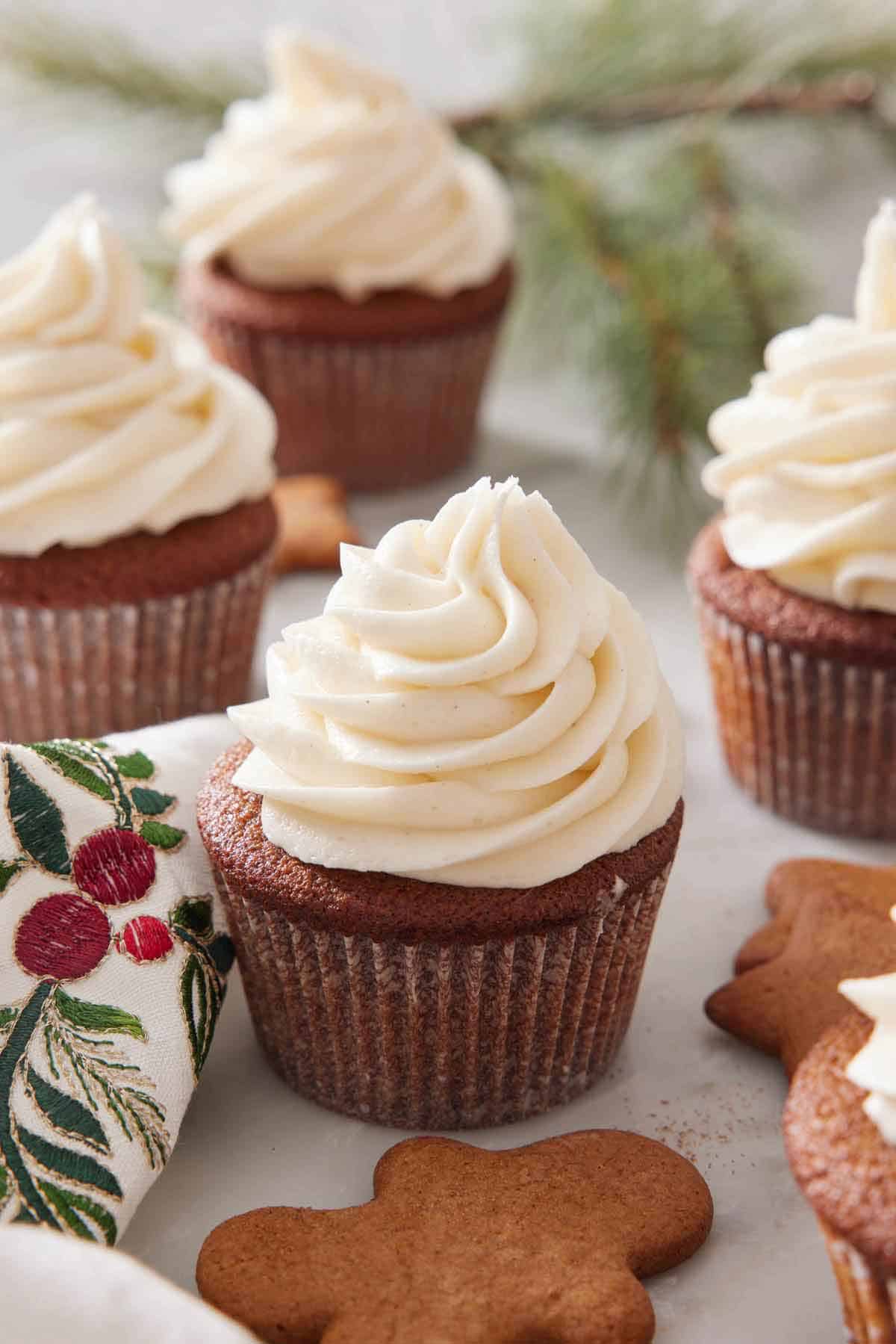 Multiple gingerbread cupcakes with cream cheese frosting with gingerbread cookies scattered around.