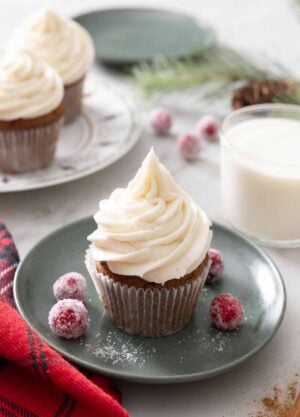 A plate with a gingerbread cupcake topped with cream cheese frosting. Two more cupcakes in the background and a glass of milk.