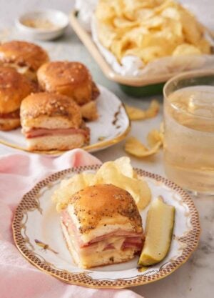 A plate with a ham and cheese slider along with chips and a pickle. More sliders, chips, and a drink in the background.