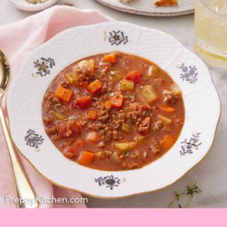 Pinterest graphic of a bowl of hamburger soup with a drink, torn bread, and pepper in the background.