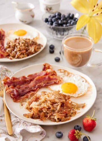 A plate with hash browns, bacon strips, and a fried egg with a cup of coffee, bowl of blueberries, and additional plate in the background.