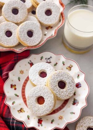 A festive plate with three linzer cookies by a mug of milk and a platter of cookies.