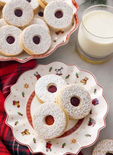 A festive plate with three linzer cookies by a mug of milk and a platter of cookies.
