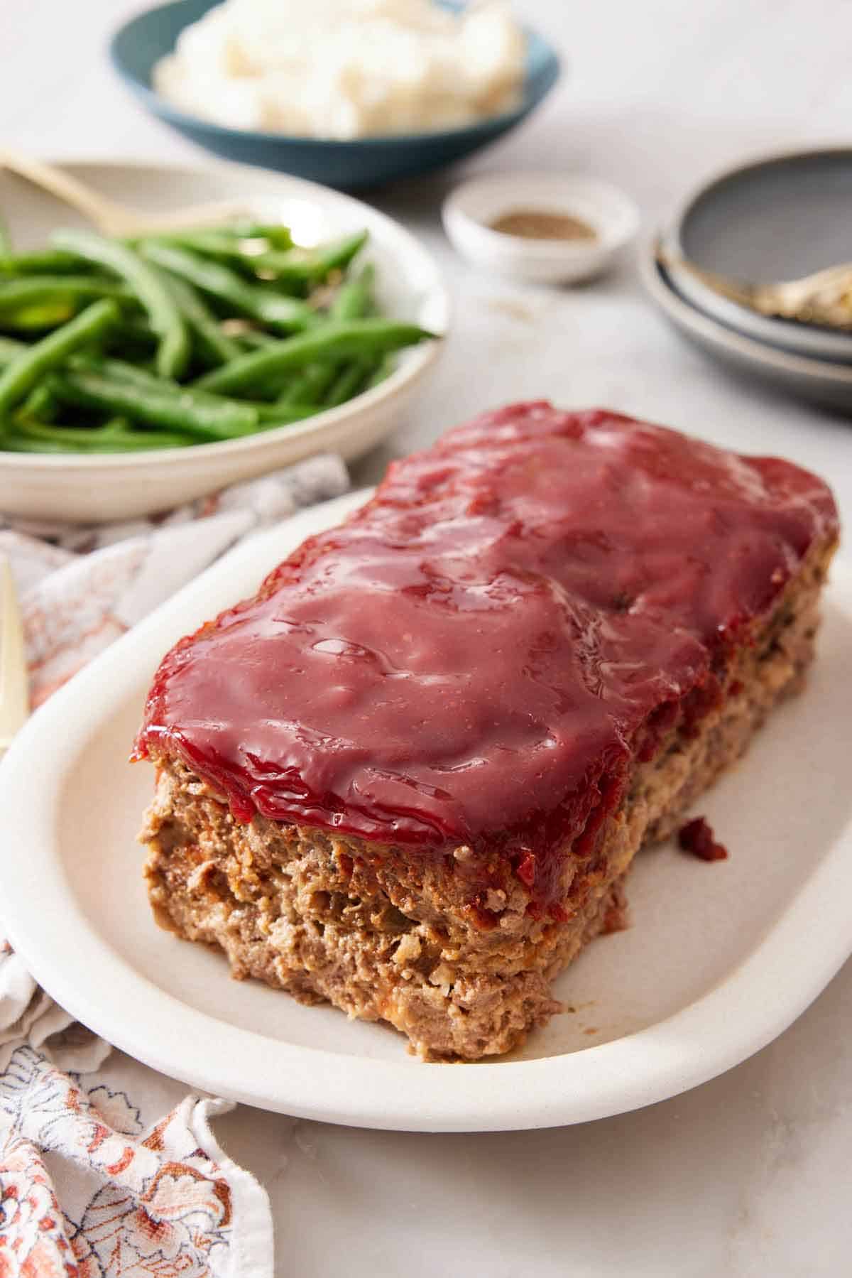 A platter of with a loaf of meatloaf with some green beans and mashed potatoes in the background.