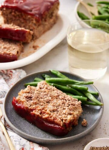 A slice of meatloaf with green beans on a plate with a glass of wine and the rest of the meatloaf in the background.